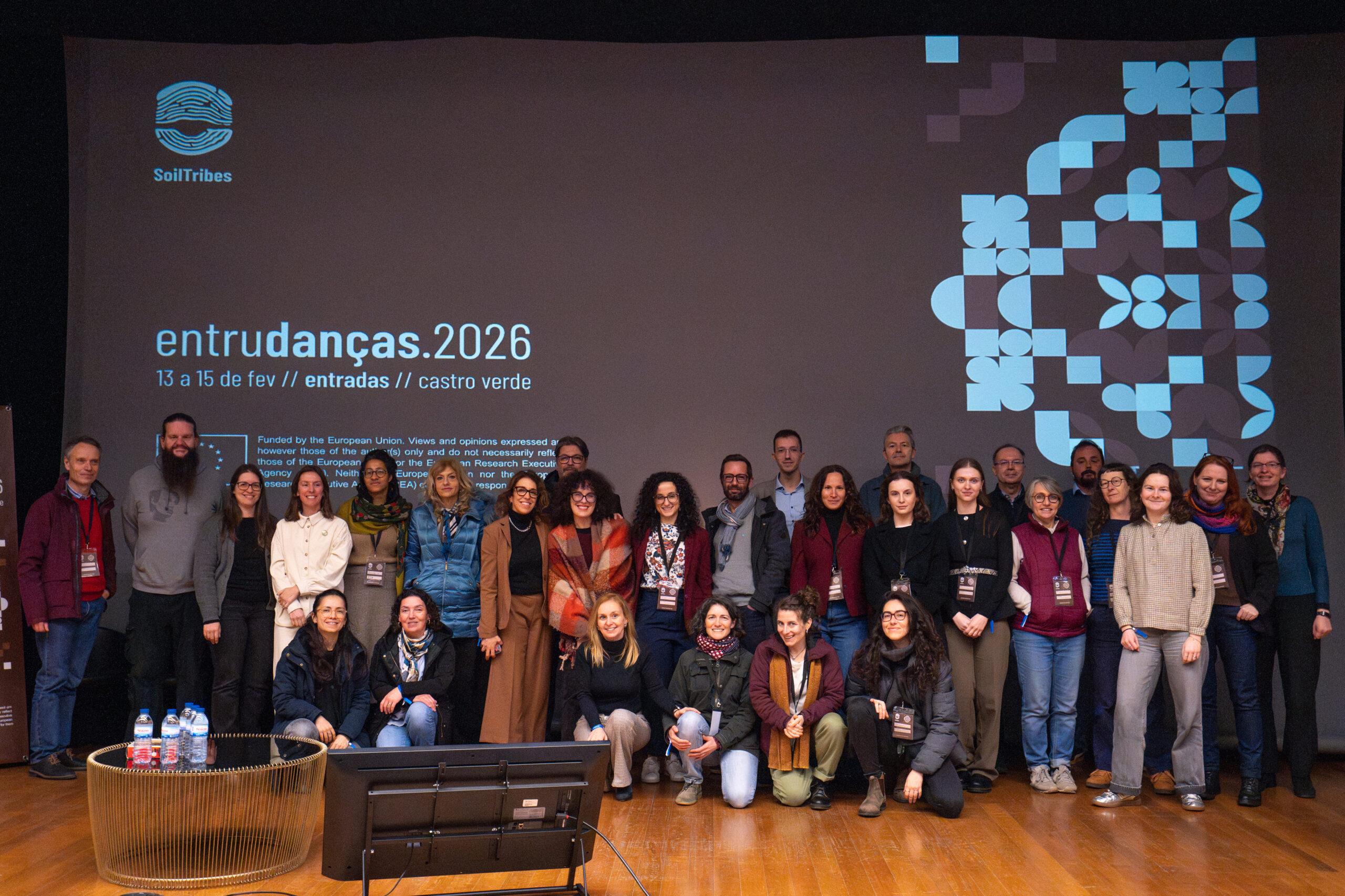 A group of SoilTribes partners stands together on a stage in front of a large screen during the consortium meeting in Beja, Portugal. The screen displays the SoilTribes logo and promotional visuals for “entrudanças 2026,” scheduled for 13–15 February in Entradas, Castro Verde