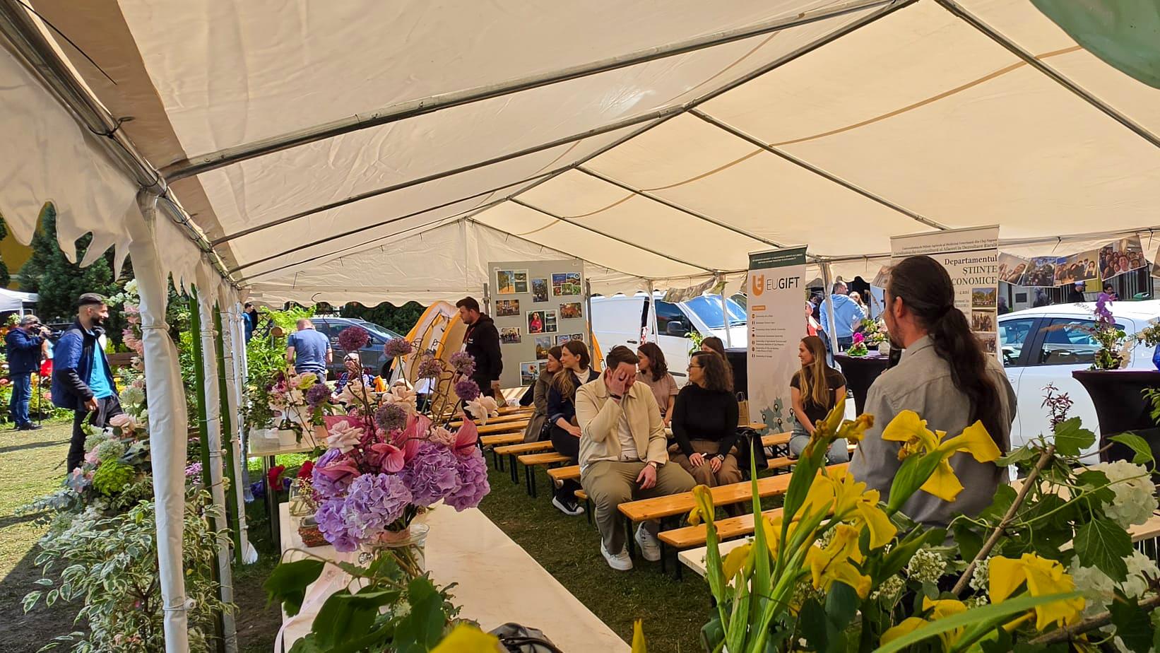 Participants attend the SoilTribes End-User Action Lab inside a dedicated tent at an outdoor eco-agricultural fair in Cluj-Napoca, surrounded by flowers and local exhibitors.