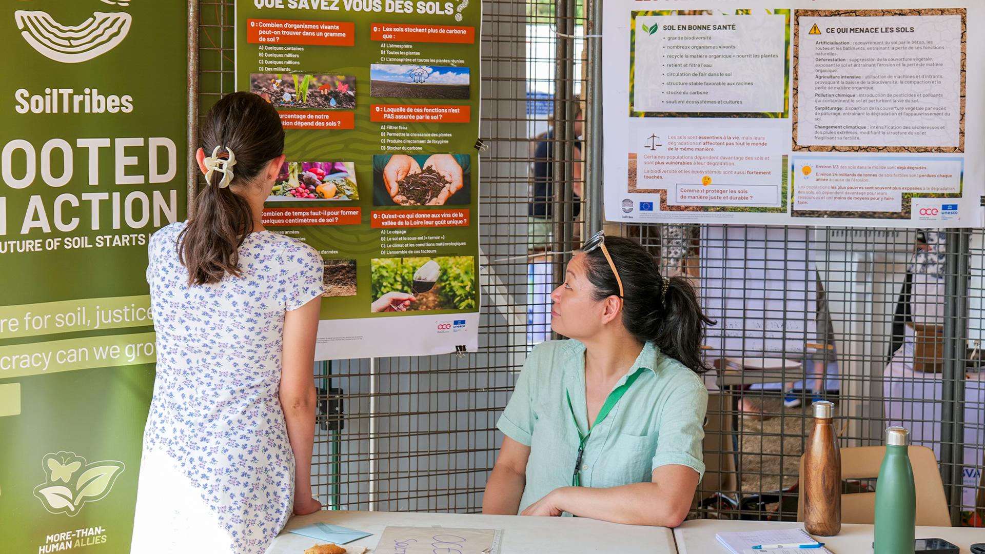 Two people at a SoilTribes interactive stand, looking at soil education posters and project materials displayed on a metal grid wall.