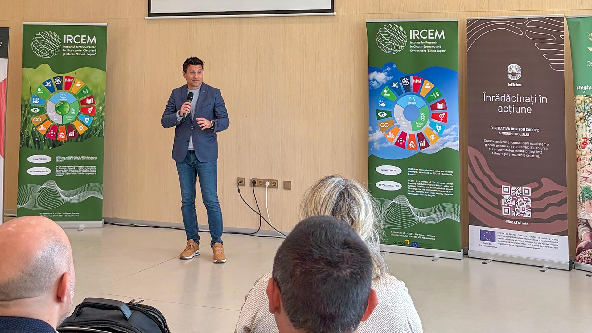 A facilitator speaks to participants during the SoilTribes Train-the-Trainers Session at IRCEM in Cluj, with SoilTribes and IRCEM banners displayed behind him.