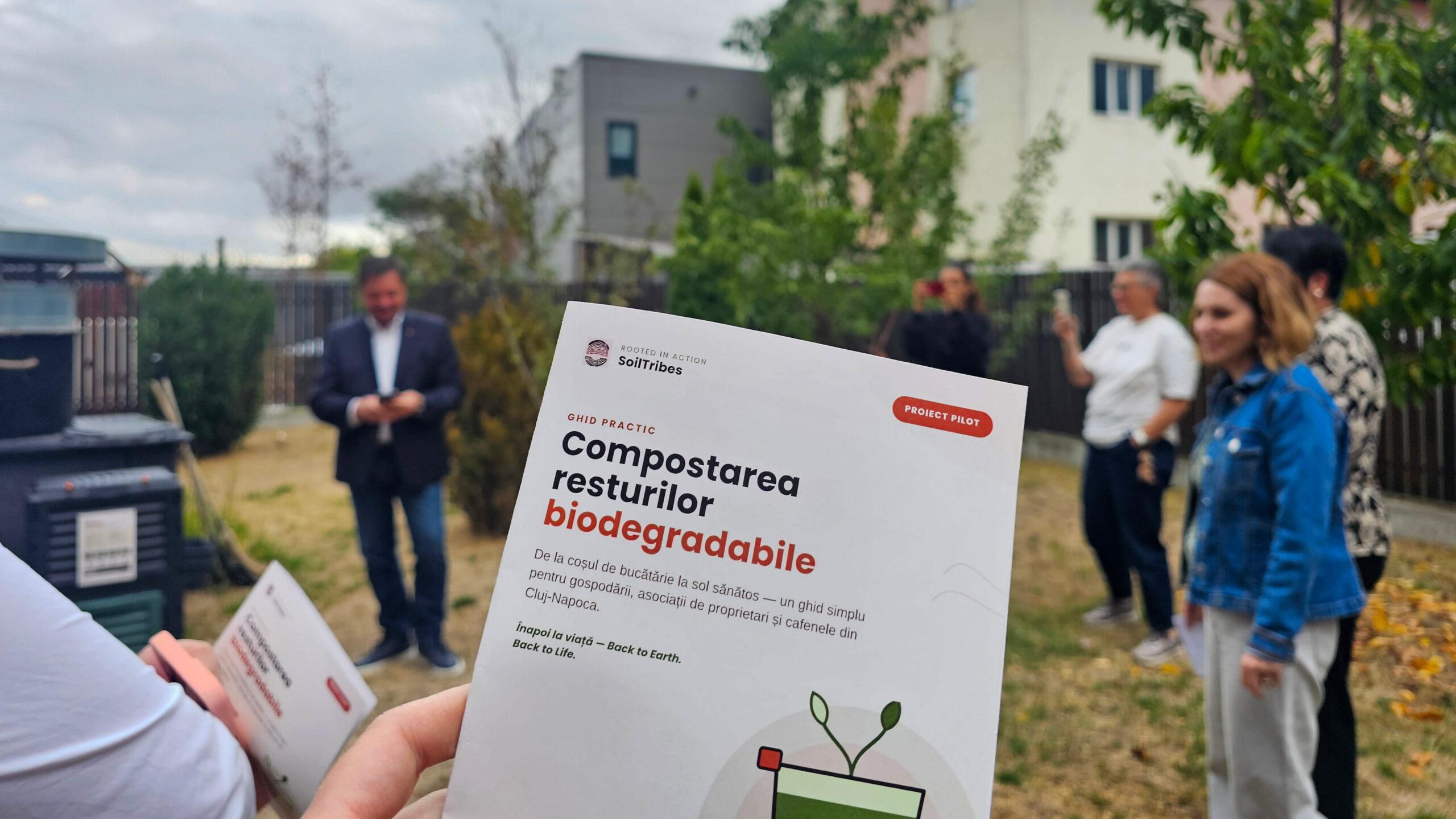A participant holds a SoilTribes practical guide on biodegradable waste composting during the urban composting pilot in Cluj-Napoca, Romania, with community members and composting bins visible in the background.
